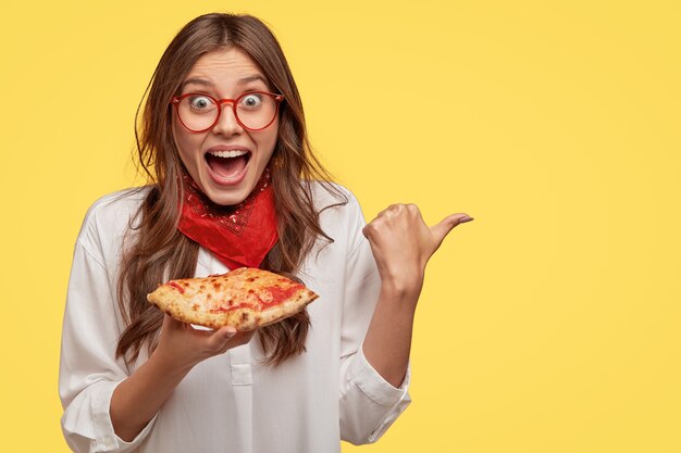 幸せな若い女性は幸せそうに見つめ、自由空間を親指で脇に向け、ピザを食べ、方向を示し、顎を落とし続け、幸せを叫び、黄色い壁に隔離されます。