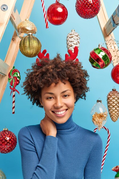 Pleased happy young woman keeps hand on neck dressed casually smiles gently has festive mood poses against blue background with new year toys hanging behind. Holidays and celebration concept