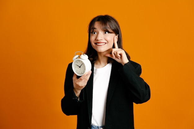 Pleased holding alarm clock young beautiful female wearing black jacket isolated on orange background