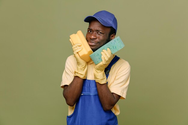 Pleased holding cleaning sponges around face young africanamerican cleaner male in uniform with gloves isolated on green background