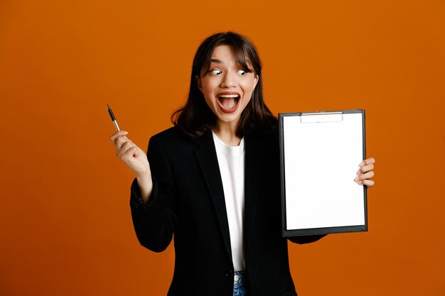 Pleased holding clipboard young beautiful female wearing black jacket isolated on orange background