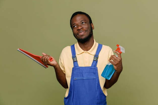 Pleased holding mop with cleaning agent young africanamerican cleaner male in uniform with gloves isolated on green background