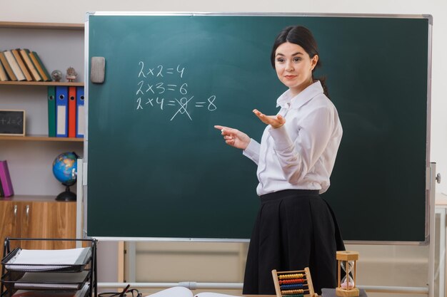 pleased holding out hand at camera young female teacher standing in front blackboard and write in classroom