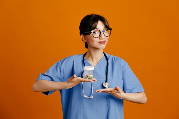 주황색 배경에 격리된 제복을 입은 젊은 여성 의사가 환약 용기를 들고 기쁘게 생각합니다.