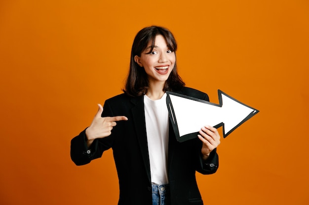 Pleased holding and points at direction mark young beautiful female wearing black jacket isolated on orange background