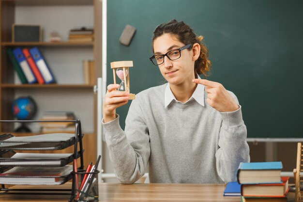 教室で学校の道具をつけて机に座って眼鏡をかけている若い男性教師が砂時計を持ってポイントすることを喜んでいます