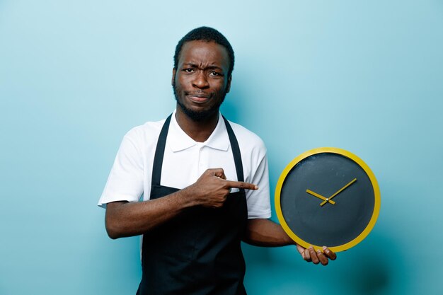 Holding felice e punti all'orologio da parete giovane barbiere afroamericano in uniforme isolato su sfondo blu