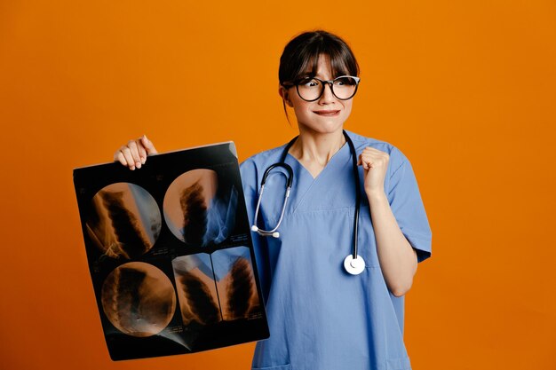 オレンジ色の背景で隔離の均一なフィス聴診器を身に着けているX線の若い女性医師を保持して満足