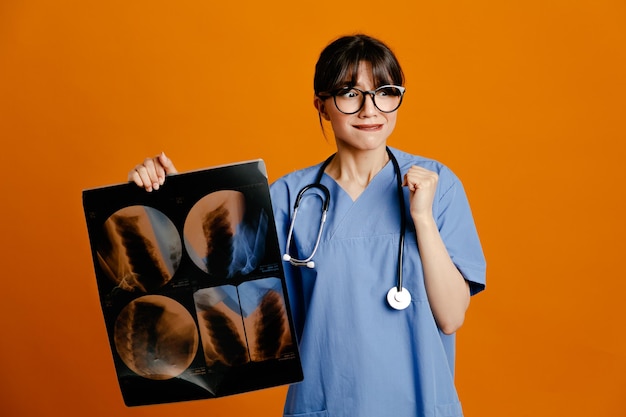 オレンジ色の背景で隔離の均一なフィス聴診器を身に着けているX線の若い女性医師を保持して満足
