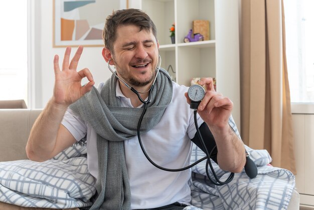 pleased ill man with scarf around neck measuring his pressure with sphygmomanometer and gesturing ok sign sitting on couch at living room