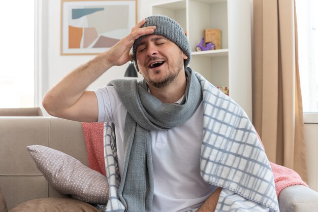 pleased ill man with scarf around neck wearing winter hat wrapped in plaid putting his hand on forehead sitting with closed eyes on couch at living room