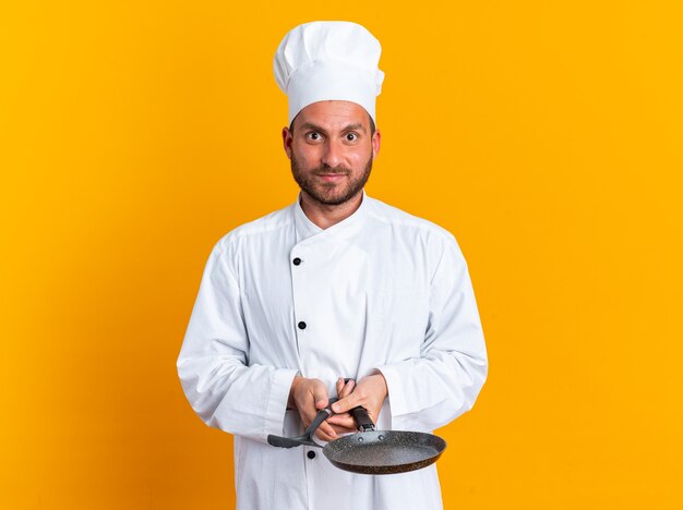 Pleased and impressed young caucasian male cook in chef uniform and cap holding spatula and frying pan looking at camera isolated on orange wall with copy space