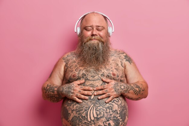 Pleased joyful bearded adult man keeps hands on big belly, enjoys awesome music in good quality wireless headphones, has problem of overweight, closes eyes and imagines something very pleasant