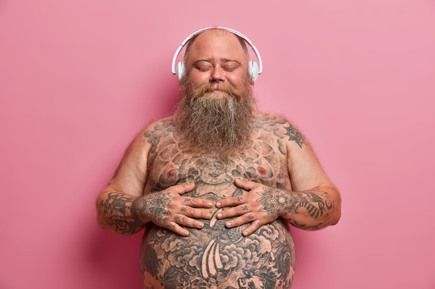 Pleased joyful bearded adult man keeps hands on big belly, enjoys awesome music in good quality wireless headphones, has problem of overweight, closes eyes and imagines something very pleasant