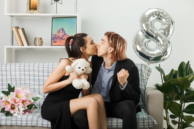 Pleased kissing each other young couple on happy women day with teddy bear sitting on sofa in living room