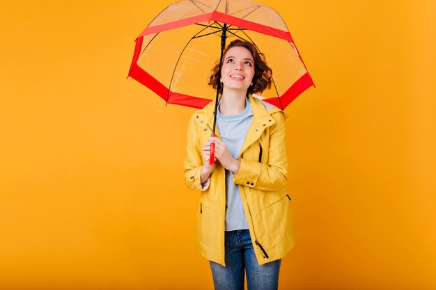 Бесплатное фото Довольная смеющаяся девушка в модном осеннем наряде, стоящая под зонтиком. крытый портрет потрясающей молодой женщины, наслаждающейся фотосессией с зонтиком.