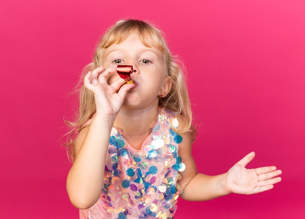 pleased little blonde girl blowing party whistle isolated on pink wall with copy space