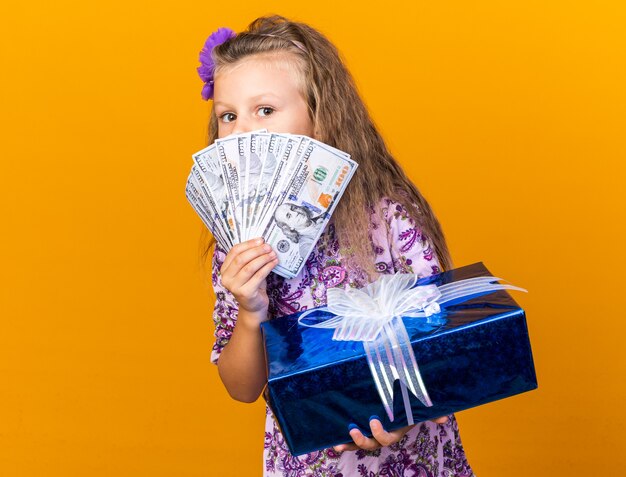 pleased little blonde girl holding gift box and money in front of her face isolated on orange wall with copy space