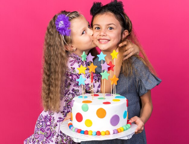 pleased little blonde girl kissing smiling little caucasian girl holding birthday cake isolated on pink wall with copy space