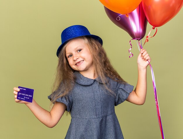 pleased little blonde girl with blue party hat holding helium balloons and credit card isolated on olive green wall with copy space