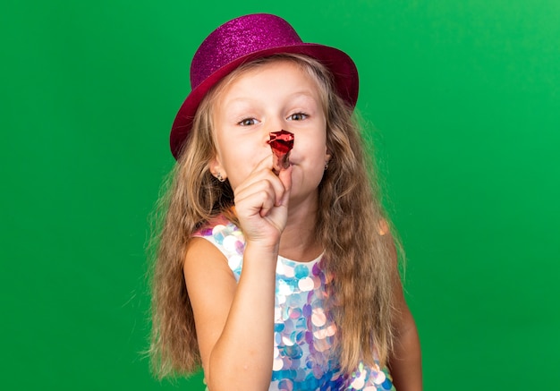 Compiaciuta bimba bionda con cappello da festa viola che soffia fischio di festa isolato sulla parete verde con spazio di copia