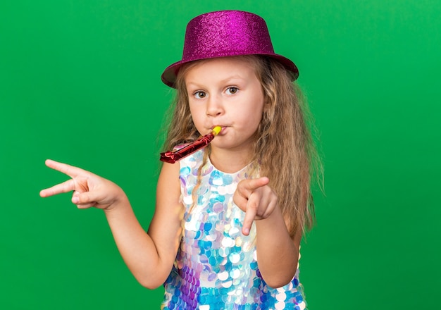 pleased little blonde girl with purple party hat blowing party whistle and pointing at sides isolated on green wall with copy space