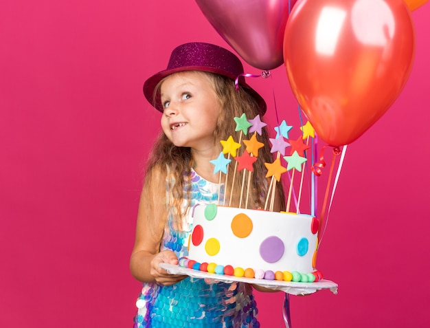 довольная маленькая блондинка в фиолетовой шляпе, держащая гелиевые шары и торт ко дню рождения, изолированную на розовой стене с копией пространства