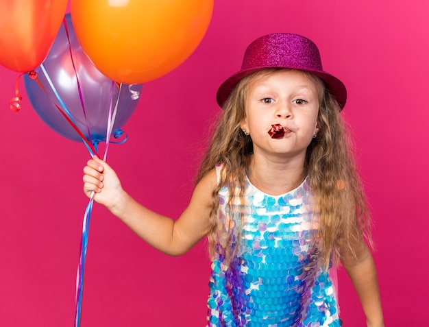 pleased little blonde girl with purple party hat holding helium balloons and blowing party whistle isolated on pink wall with copy space
