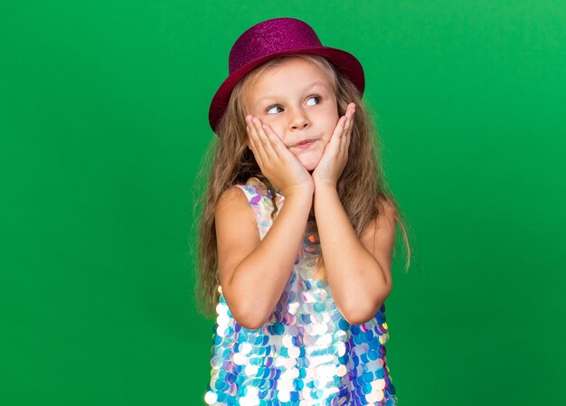 보라색 파티 모자와 함께 기쁘게 작은 금발 소녀 얼굴에 손을 넣고 복사 공간이 녹색 벽에 고립 된 측면에서 보인다