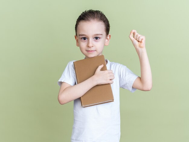 Pleased little boy holding book looking at camera doing ok sign isolated on olive green wall with copy space