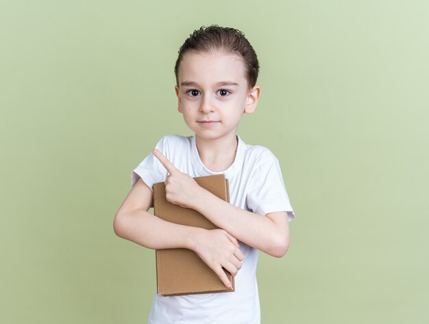 Pleased little boy holding book looking at camera pointing up isolated on olive green wall with copy space