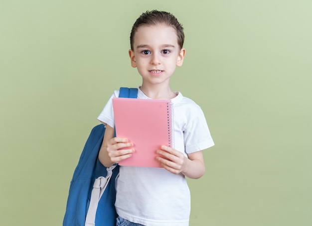 Pleased little boy wearing backpack looking at camera showing note pad isolated on olive green wall with copy space