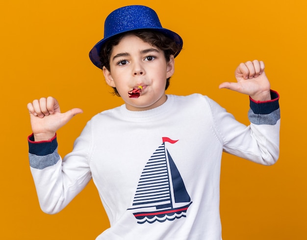 Pleased little boy wearing blue party hat blowing party whistle points at himself 