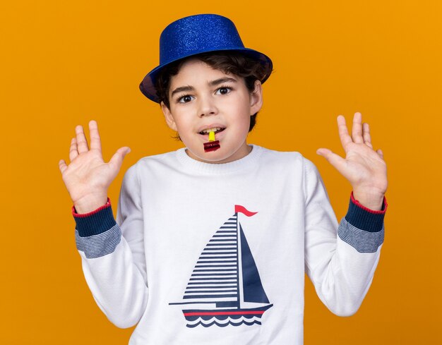 Pleased little boy wearing blue party hat blowing party whistle spreading hands 