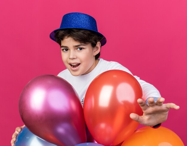 Pleased little boy wearing blue party hat standing behind balloons holding out hand at camera 