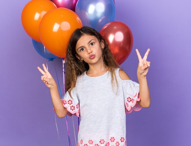 Foto gratuita contenta piccola ragazza caucasica che gesturing segno di vittoria in piedi di fronte a palloncini di elio isolati sulla parete viola con spazio di copia