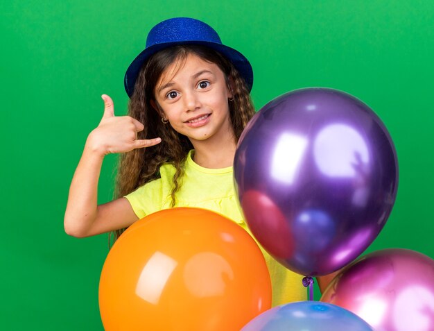 ヘリウム気球を保持し、コピースペースで緑の壁に分離された緩いジェスチャーをぶら下げて青いパーティーハットで喜んでいる小さな白人の女の子