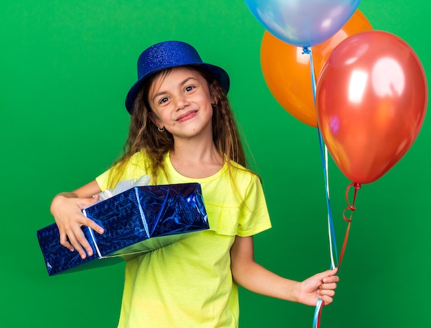 pleased little caucasian girl with blue party hat holding helium balloons and gift box isolated on green wall with copy space