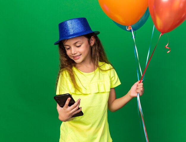 pleased little caucasian girl with blue party hat holding helium balloons and looking at phone isolated on green wall with copy space