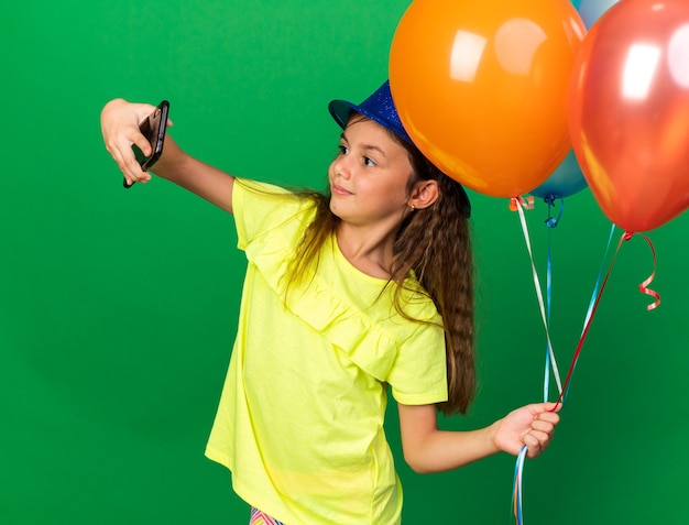Довольная маленькая кавказская девушка в синей партийной шляпе держит гелиевые шары и делает селфи на телефоне, изолированном на зеленой стене с копией пространства