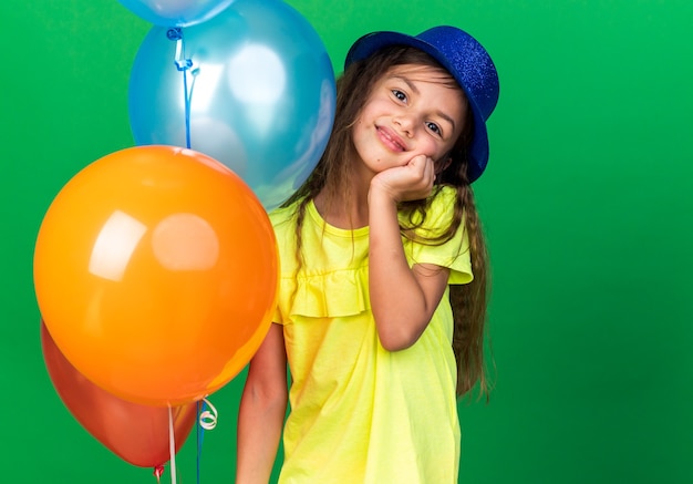 pleased little caucasian girl with blue party hat putting hand on face and holding helium balloons isolated on green wall with copy space