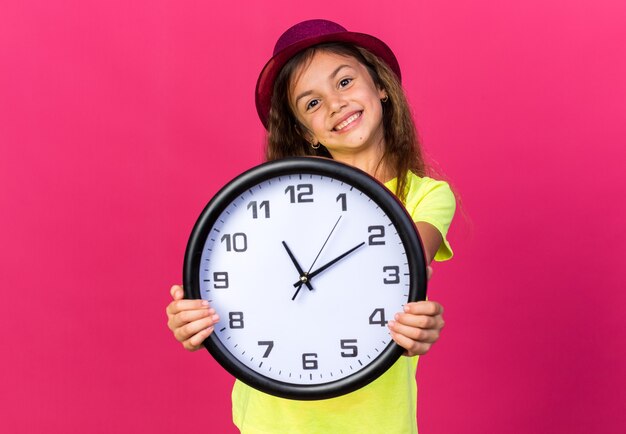 pleased little caucasian girl with purple party hat holding clock isolated on pink wall with copy space