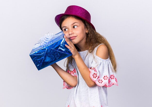 pleased little caucasian girl with purple party hat holding gift box isolated on white wall with copy space