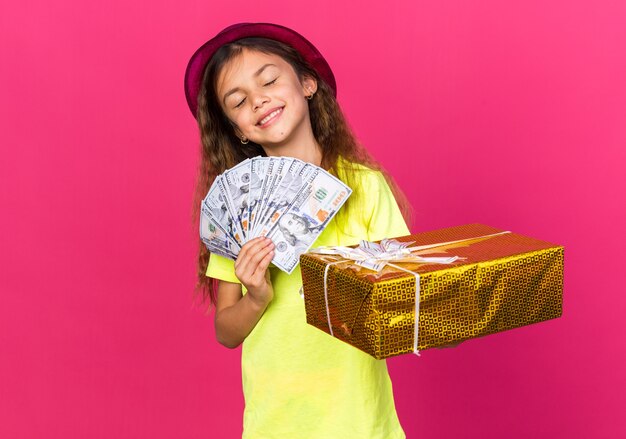 pleased little caucasian girl with purple party hat holding gift box and money isolated on pink wall with copy space