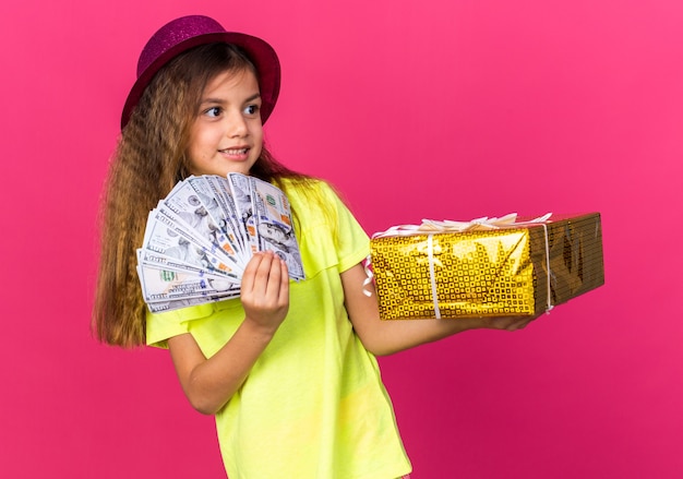 pleased little caucasian girl with purple party hat holding gift box and money looking at side isolated on pink wall with copy space