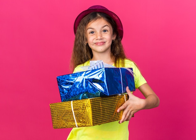 복사 공간 분홍색 벽에 고립 된 선물 상자를 들고 보라색 파티 모자와 함께 기쁘게 어린 백인 소녀