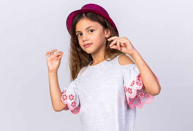pleased little caucasian girl with purple party hat pretending to hold something isolated on white wall with copy space