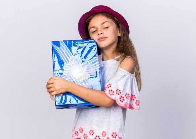 pleased little caucasian girl with purple party hat standing with closed eyes holding gift box isolated on white wall with copy space