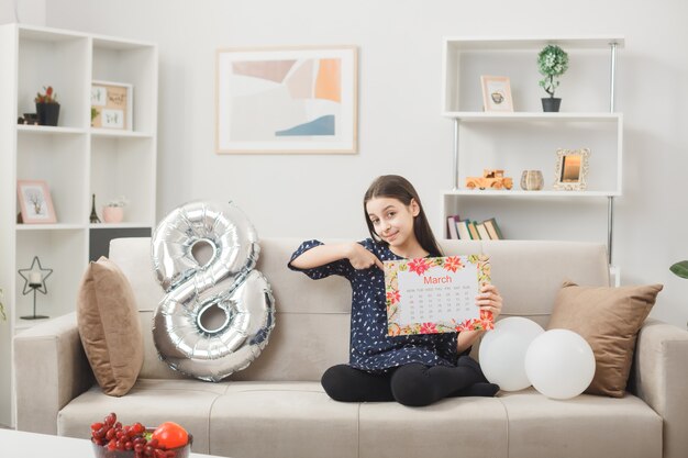 Pleased little girl on happy women's day holding and points at calendar sitting on sofa in living room