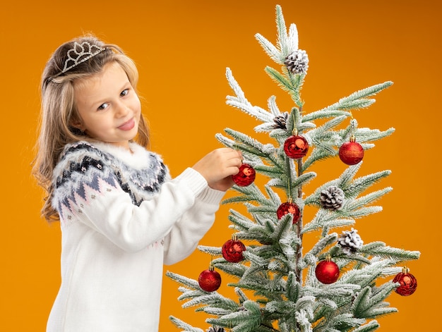 Довольная маленькая девочка, стоящая рядом с елкой в тиаре с гирляндой на шее, вешает игрушку на елку, изолированную на оранжевом фоне